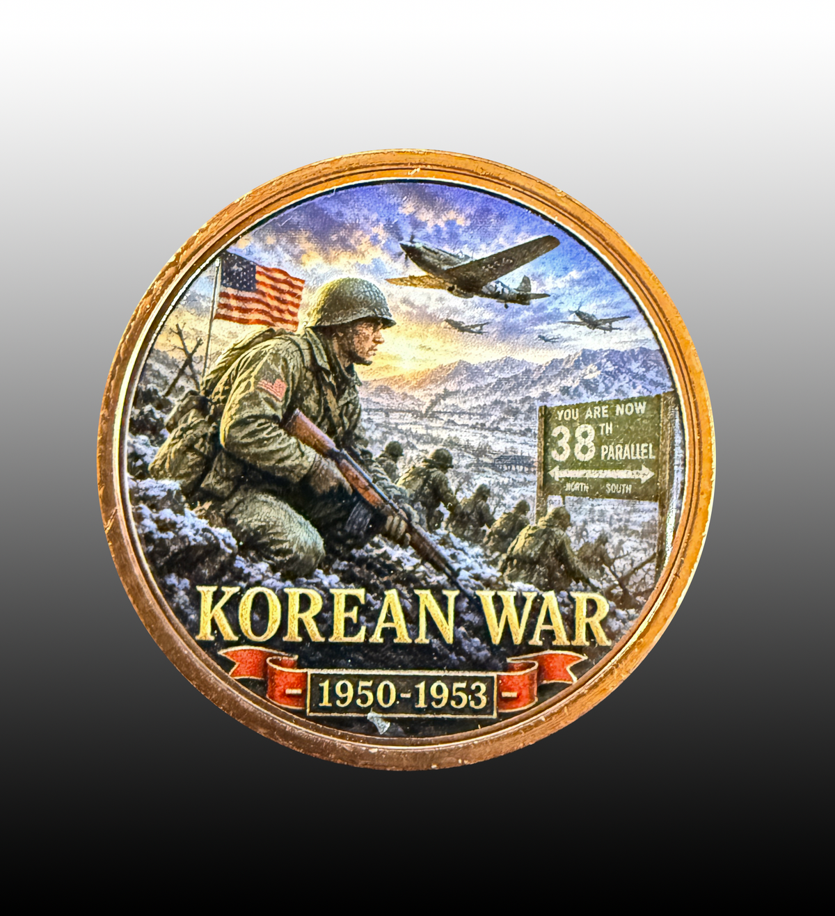 A circular commemorative coin or medallion featuring a Korean War scene with a soldier in the snow, military planes, mountains, and the text 'KOREAN WAR 1950-1953' on a gold-toned rim against a gray-to-black gradient background.